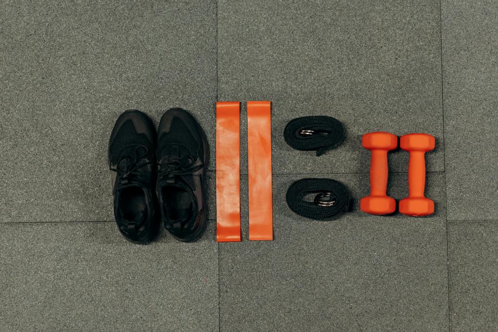 Workout equipment neatly arranged on gray floor: black athletic shoes, orange resistance bands, fitness tracker or heartrate monitor, and orange dumbbells, representing a complete home exercise setup