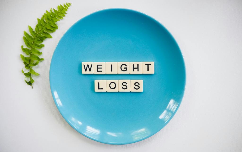 Bright turquoise plate with letter tiles spelling 'WEIGHT LOSS' arranged in the center, accompanied by a small green fern leaf to the left against a white background, symbolizing healthy eating and weight management
