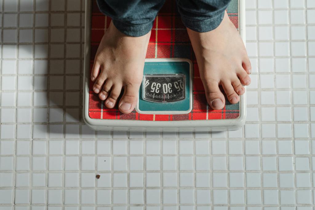 A person is standing on a bathroom scale with a red and white checkered design. The scale displays a weight of 56.06, though the unit is not specified. The floor is tiled with small white squares, and the person is wearing blue jeans.